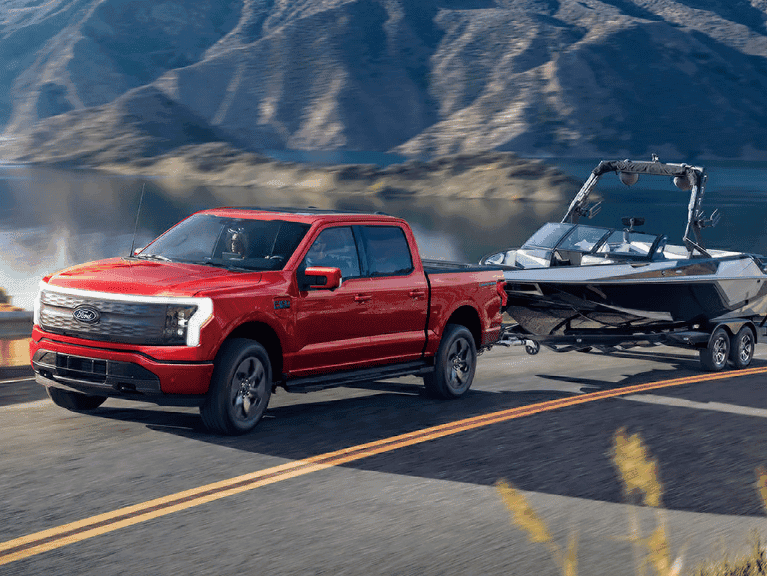 A 2025 Ford F-150® Lightning® pickup pulling a boat on a trailer down a highway near a mountain lake
