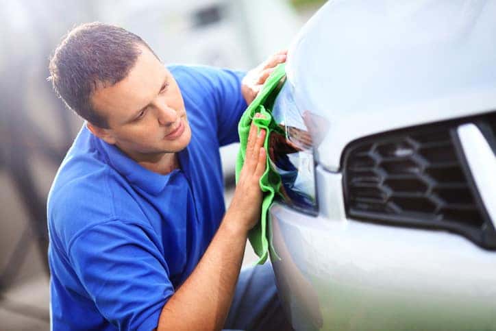 Mid-aged man polishing headlight on his silver metallic colored vehicle with green cloth at parking lot. He's kneeling next to his car.He has brown hair and wearing blue polo shirt.