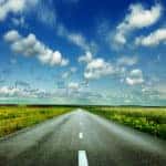 Long paved road with a white dotted line down the middle and grass on either side below a blue sky with white fluffy clouds in it