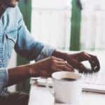 Man in a blue shirt sitting at a white table with a white cup of coffee, using a laptop