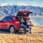 Red 2020 Ford Escape sitting next to a body of water and snowy mountains with the trunk open, two women sitting on the back, and a woman fishing in the background