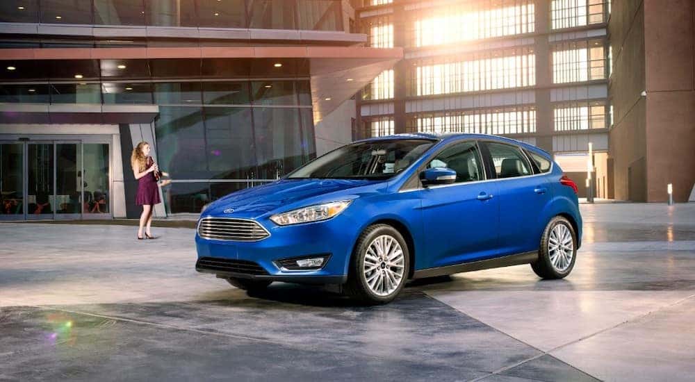 Woman in a purple dress looking at a blue 2018 Ford Focus hatchback parked in a grey and brown showroom