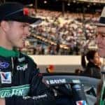 Jack Roush wearing a hat standing next to NASCAR driver at a stadium with an audience in the background