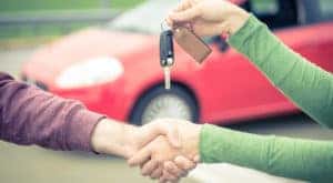 Woman in a green shirt shaking hands with a man in a purple shirt while handing him a car key in front of a red vehicle after researching used car values.