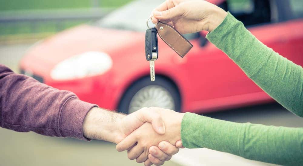 Woman in a green shirt shaking hands with a man in a purple shirt while handing him a car key in front of a red vehicle after researching used car values.