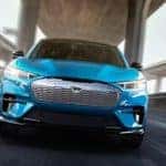 A blue 2020 Ford Mustang Mach-E is facing forward while driving under a concrete bridge.