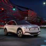 White 2021 Mustang Mach-E electric vehicle parked in an empty lot in front of a tall red, wavy fence