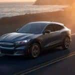 A grey 2021 Ford Mustang Mach-E is driving past the ocean at sunset.