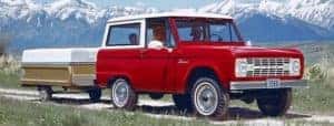A red 1966 Ford Bronco is towing a camper in front of snow-covered mountain.