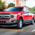 A red 2020 Ford F-150 with a Powerstroke diesel engine is towing a trailer full of trees on a city street.