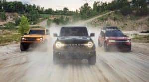 A yellow 2021 Ford Bronco, a black Bronco and a red Bronco Sport are shown from the front driving on a dirt road after leaving a NY Ford Bronco dealer.
