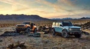 A yellow 2021 Ford Bronco 2 door with no doors and a blue Bronco 4 door are parked on rocks in a desert in front of distant mountains.