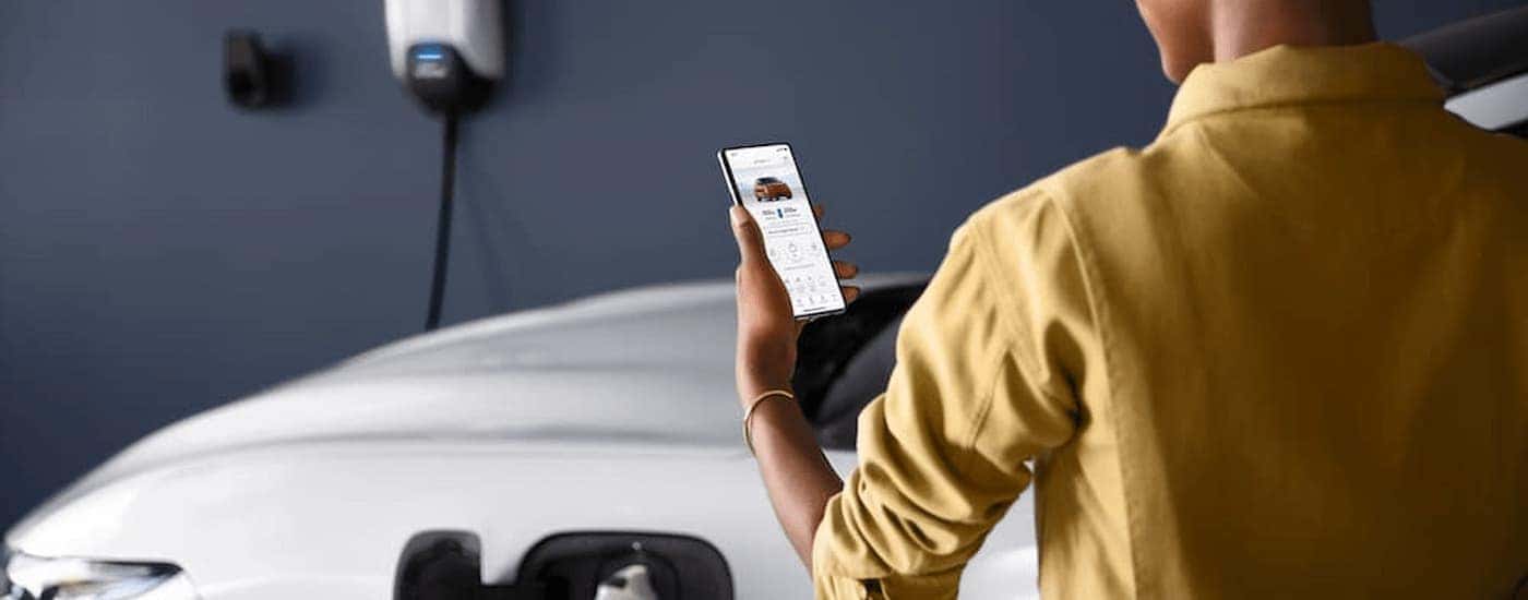 A woman is shown looking at her phone next to a charging white 2022 Ford Mustang Mach-E.