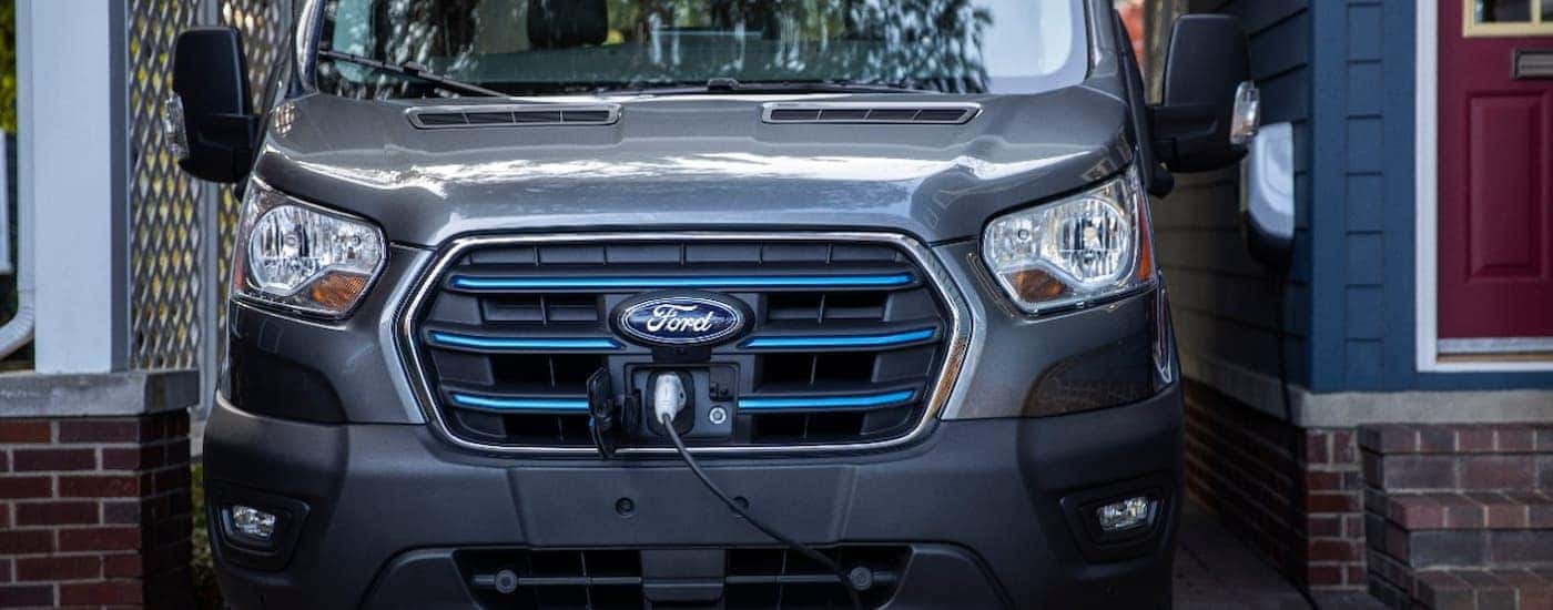 The front of a grey and blue 2023 Ford E-Transit is shown charging at a Ford electric car dealership.