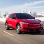 A red 2021 Ford Mustang Mach-E is driving away from a city after a Ford electric car dealership.