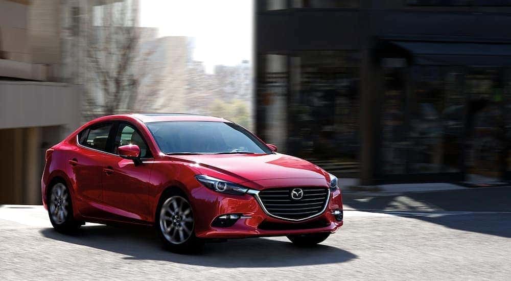 A red 2018 Maxda3 Sedan is driving through a city aftr leaving a used Mazda dealership.