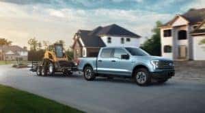 A light blue 2022 Ford F-150 Lightning Pro is shown towing construction equipment through a suburban neighborhood.