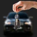 A hand is shown holding a set of car keys in front of a black sedan.