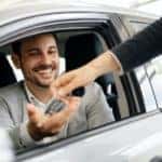 A man is shown smiling from the drivers seat of a car as a salesperson passes a set of keys.