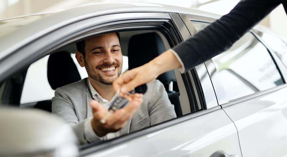 A man is shown smiling from the drivers seat of a car as a salesperson passes a set of keys.
