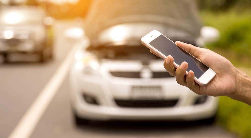 A hand is shown holding a cellphone in front of a car with the hood open.