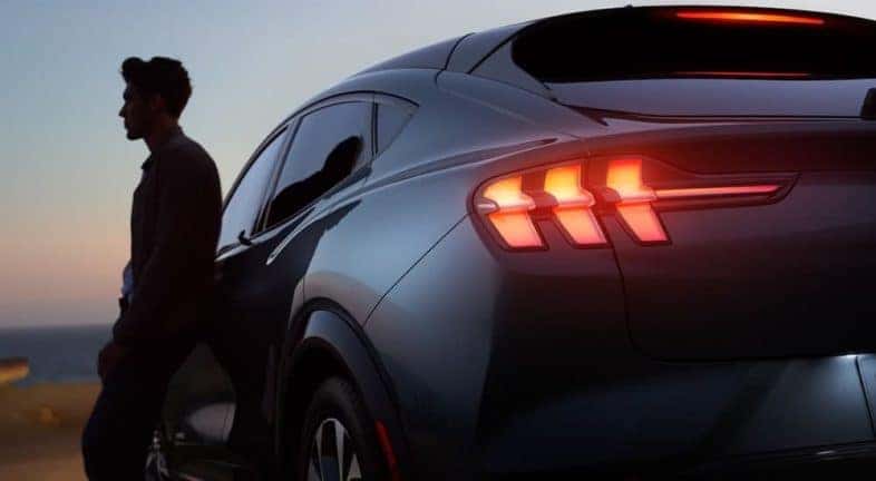A person is shown in silhouette leaning against a 2021 Ford Mustang Mach-E with illuminated taillights.