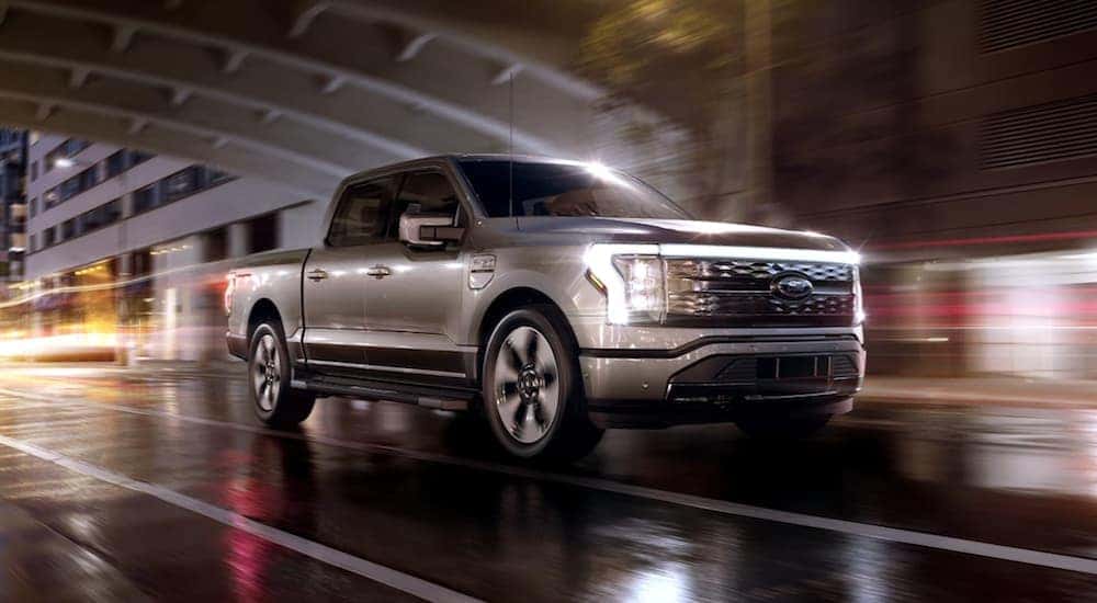 A silver 2024 Ford F-150 Lightning is shown driving through a city at night after leaving a Troy Ford dealership.