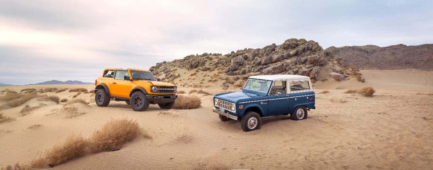 An orange 2021 Ford Bronco and a blue 1970s Ford Bronco are shown parked in a desert.