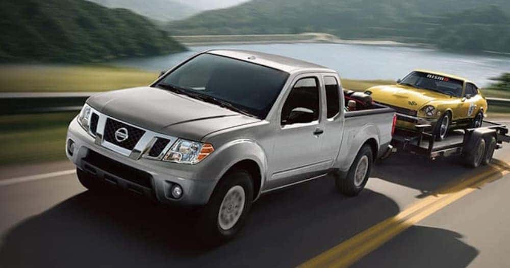 A grey 2018 Nissan Frontier is shown towing a Datsun 240z on a trailer past a body of water.