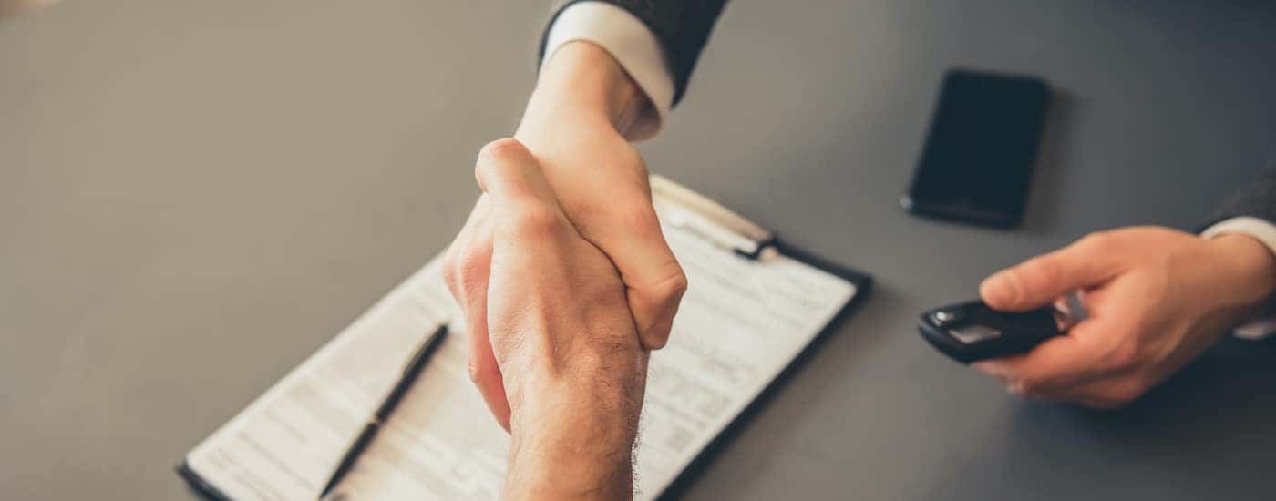 A customer is shown shaking hands with a salesperson over a clipboard with paperwork.