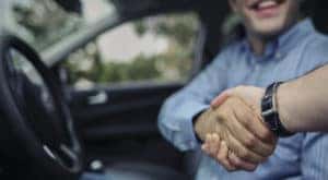 A person is shown sitting in the front seat of a car and shaking hands with a salesperson.