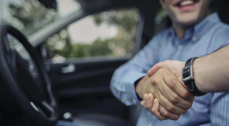 A person is shown sitting in the front seat of a car and shaking hands with a salesperson.
