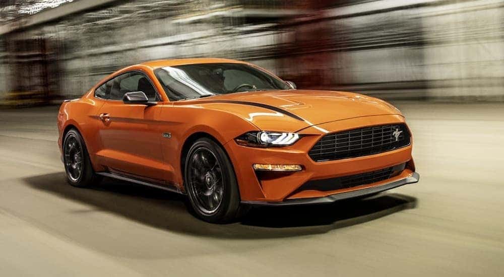 An orange 2021 Ford Mustang Ecoboost HPP is shown from the front at an angle while driving after leaving a dealership that advertises certified Ford for sale.
