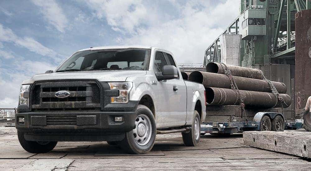 A silver 2015 Ford F-150 XL is shown towing a trailer of large pipes.