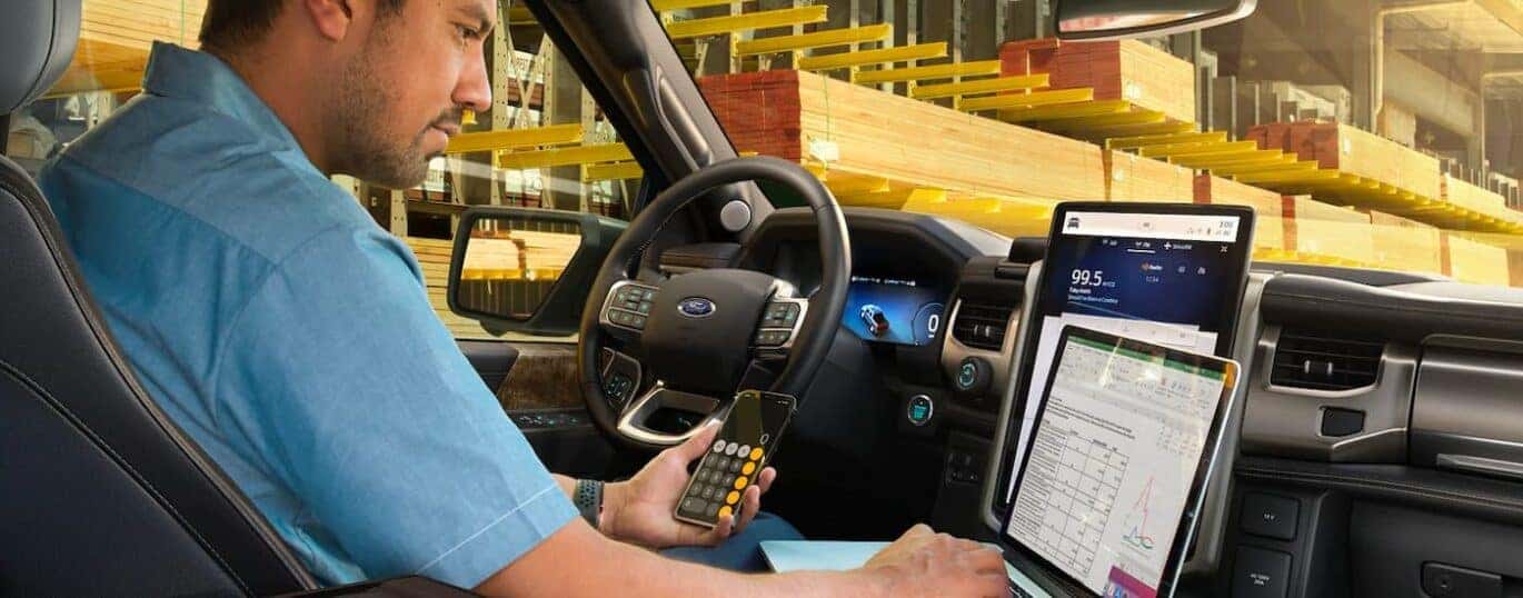 A person is shown in a 2023 Ford F-150 Lightning using a phone and laptop.