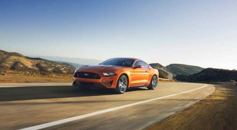 An orange 2018 Ford Mustang GT is shown driving on an open road after leaving a used car dealer near Troy.