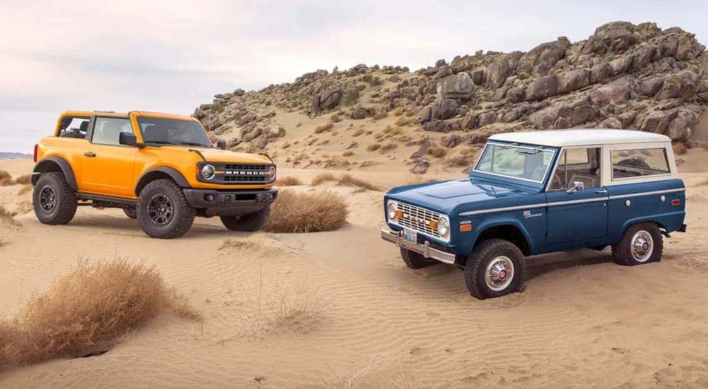 A yellow 2021 Ford Bronco and a blue 1966 Ford Bronco parked in sand outside a used Ford dealership near Halfmoon.