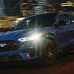 A dark blue 2024 Ford Mustang Mach-E driving past city lights at night.