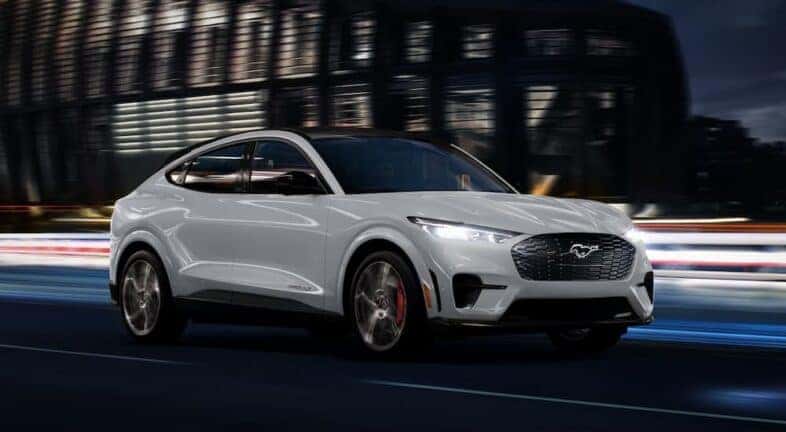 A white 2023 Ford Mustang Mach-E for sale is driving on a city street at night.