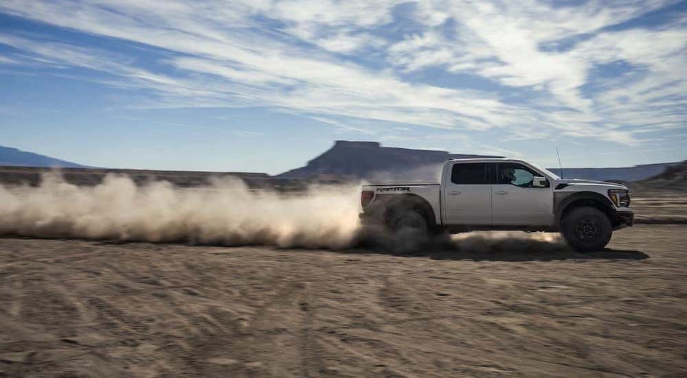 A white 2024 Ford F-150 Raptor is shown driving fast off-road.