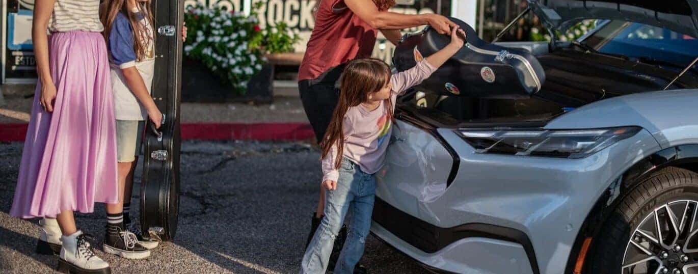 Musical instruments are shown being placed into the front trunk of a blue 2024 Ford Mustang Mach-E.
