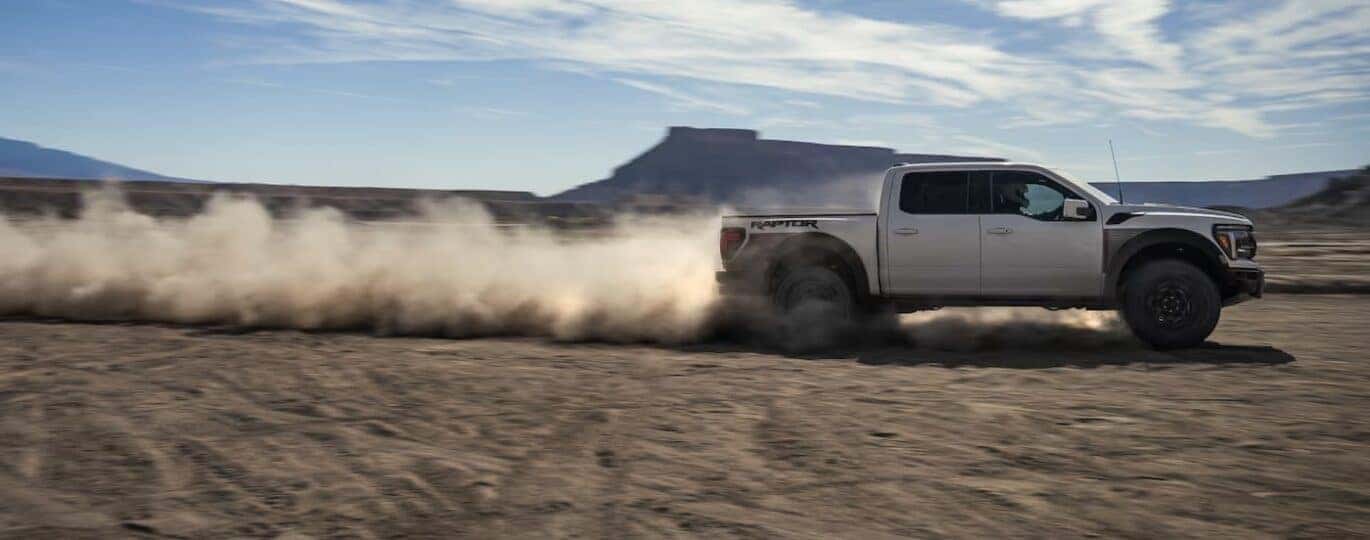 A white 2025 Ford F-150 Raptor is shown driving off-road.