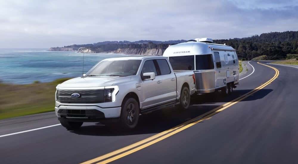 White 2023 Ford F-150 Lightning towing a camper around a bend from a dealer with used cars for sale near East Greenbush.