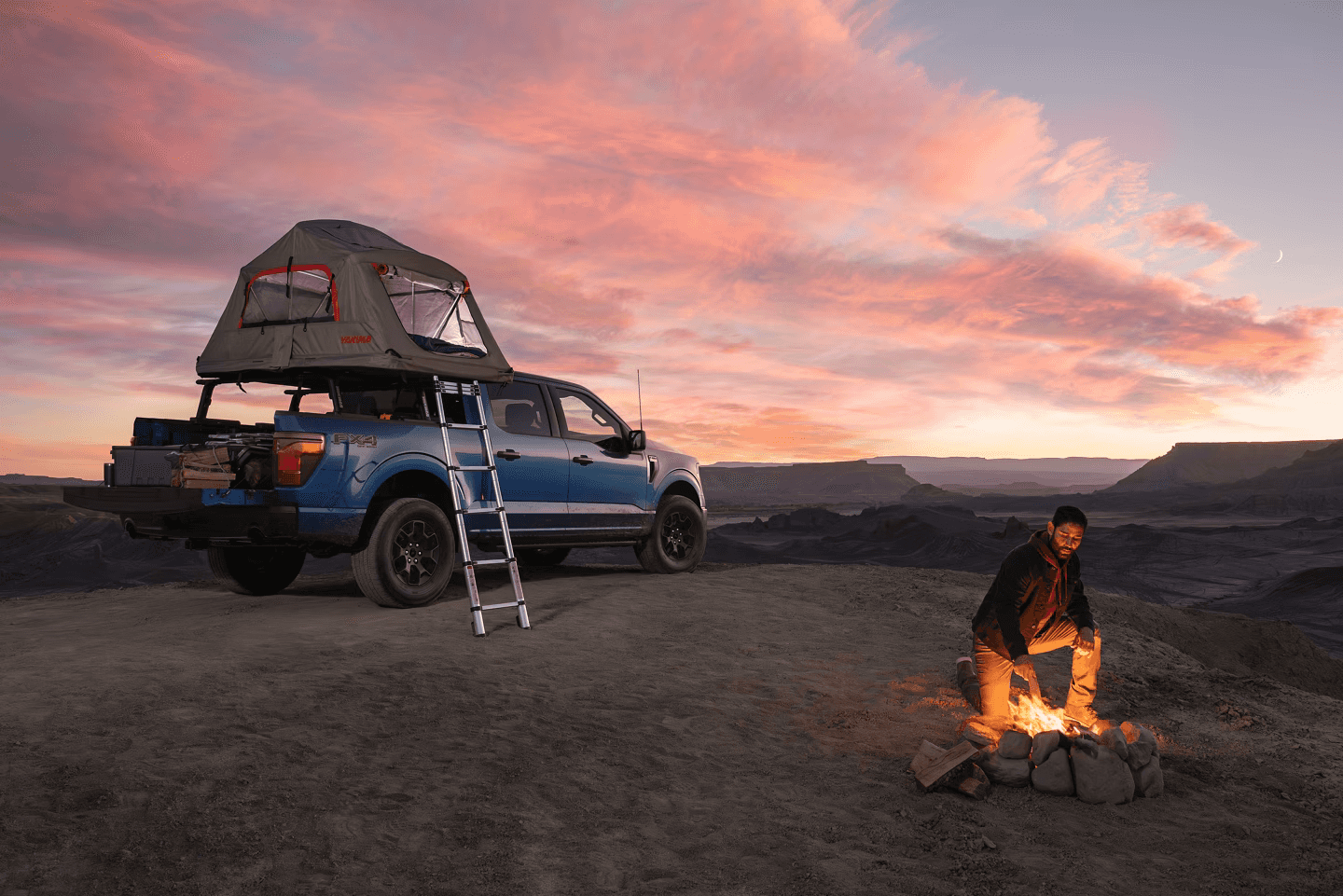 2025 Ford F-150 parked at a scenic overlook with a rooftop tent and gear, as a camper tends a fire under a vibrant sunset sky.