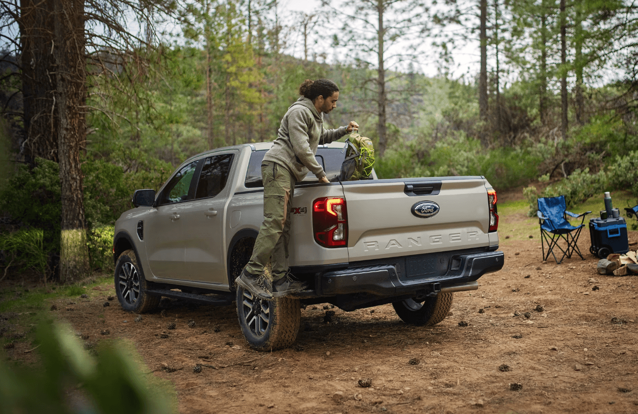 Man loading gear into a beige 2025 Ford Ranger at a forest campsite.