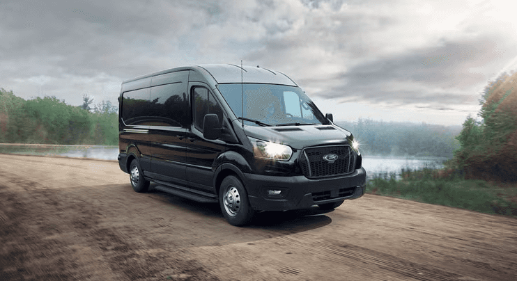 2025 Ford Transit Cargo Van in black driving on a gravel road by a lake.