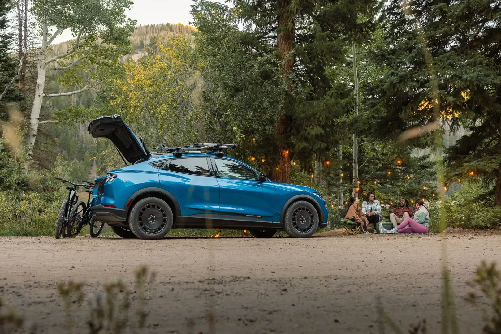Blue 2027 Ford Mustang Mach-E Trail parked at a wooded campsite with its rear hatch open, mountain bikes beside it, and four people gathered beneath string lights.