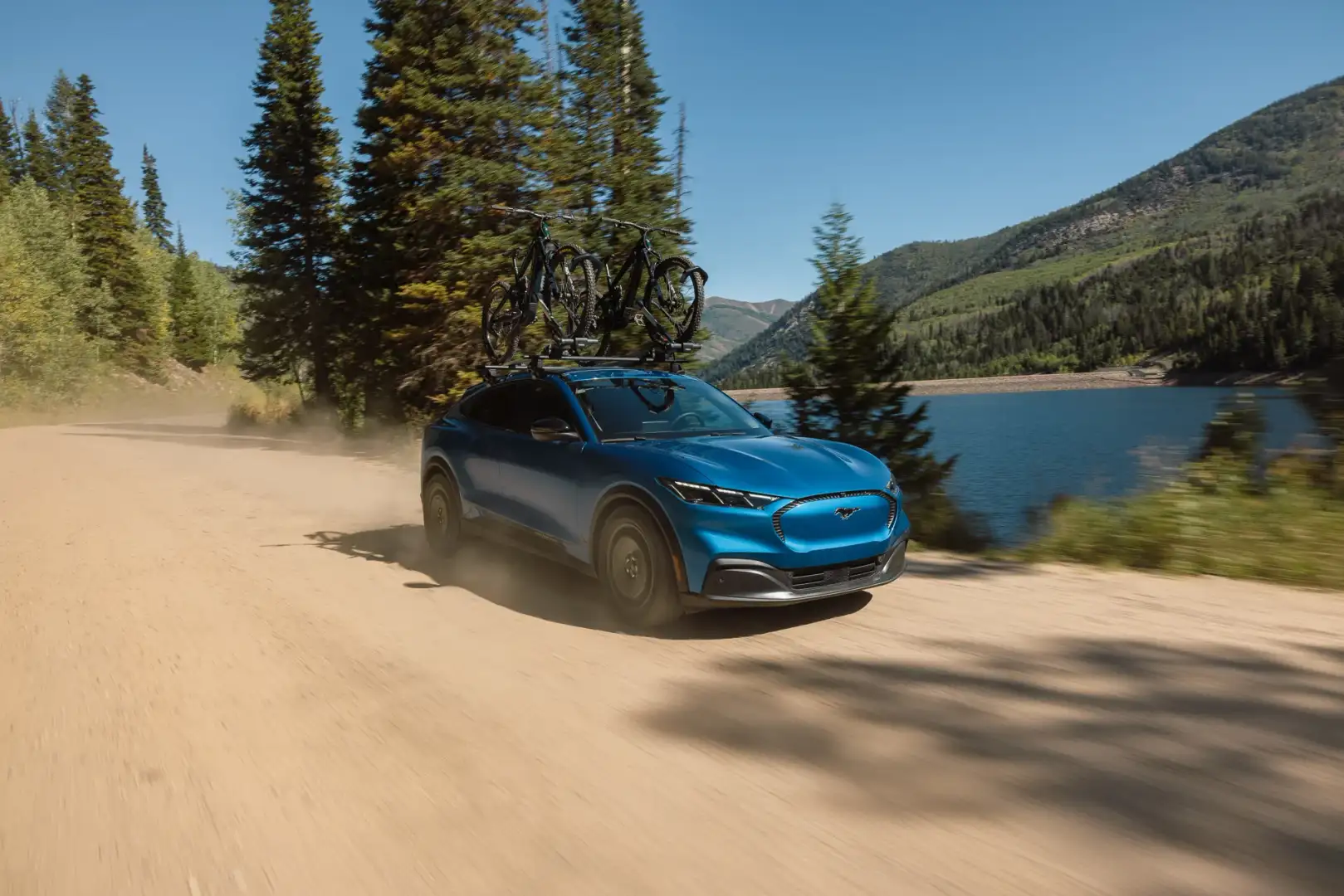 Blue 2027 Ford Mustang Mach-E Trail carrying two mountain bikes on a roof rack while driving along a dusty road beside a mountain lake.