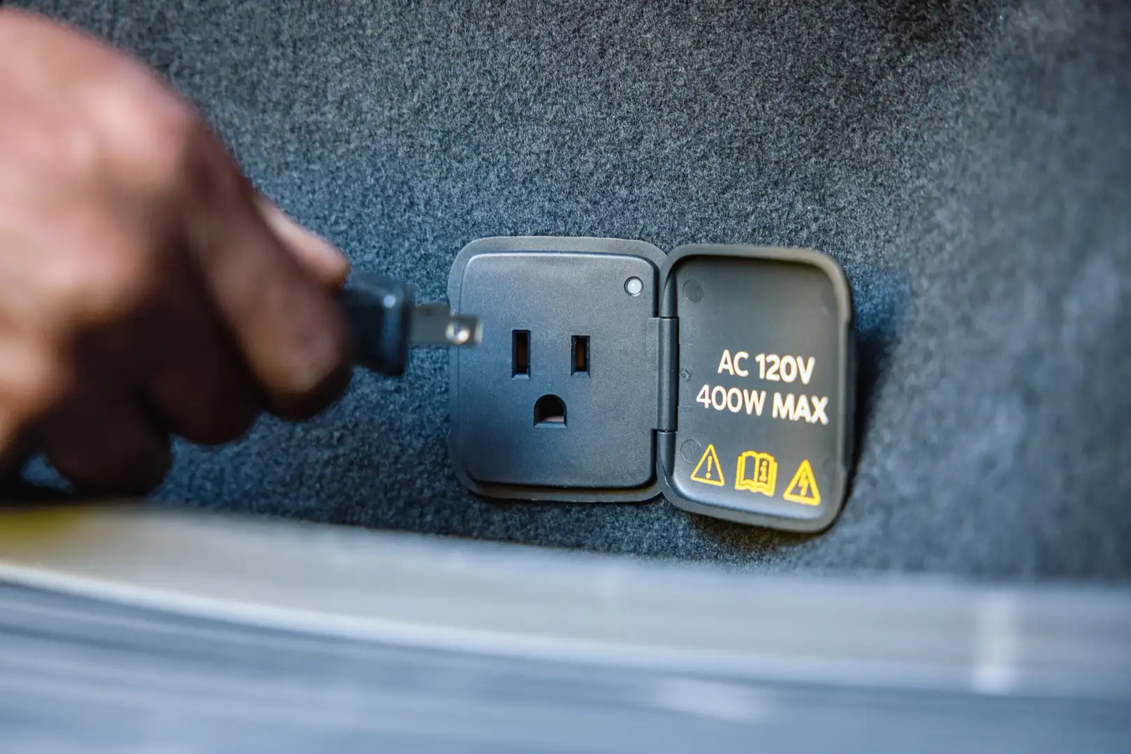 Hand holding a plug beside the 2027 Ford Mustang Mach-E Trail’s cargo-area Pro Power Onboard outlet, labeled “AC 120V, 400W Max.”