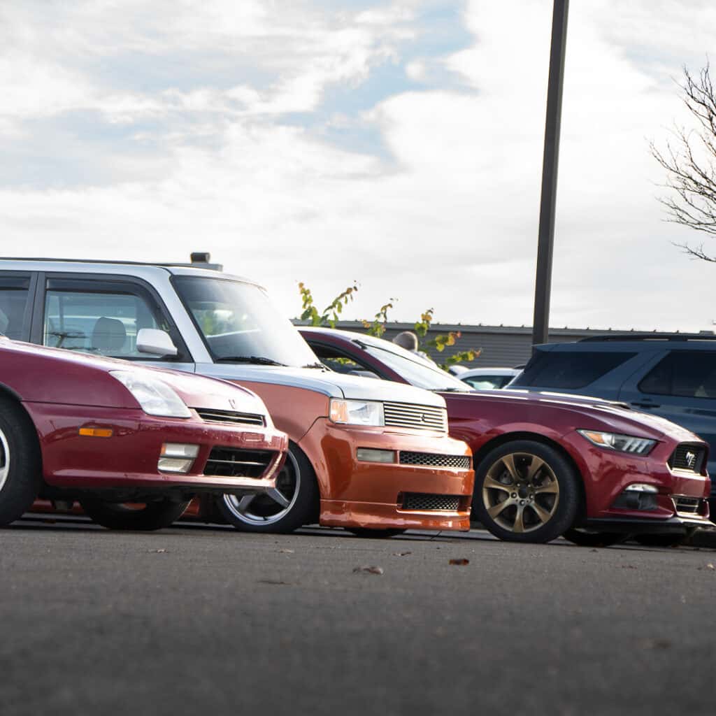 Low-angle view of a red Honda Prelude, a customized orange-and-white Scion xB, and a red Ford Mustang parked side by side at Ford of Cookeville Cars & Coffee.